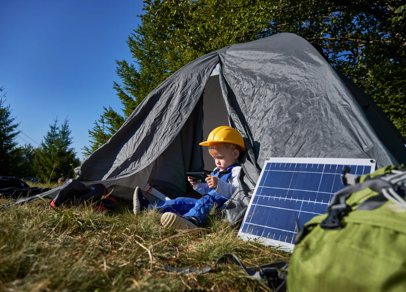 Child in a yellow construction helmet sitting in a tent with a portable solar panel propped up next to him in a sunny outdoor camp